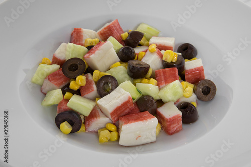 Spring salad. Closeup view of a colorful fresh salad served on a white bowl on the marble table. The salad contains sliced cucumber, crab sticks, corn grains and black olives.