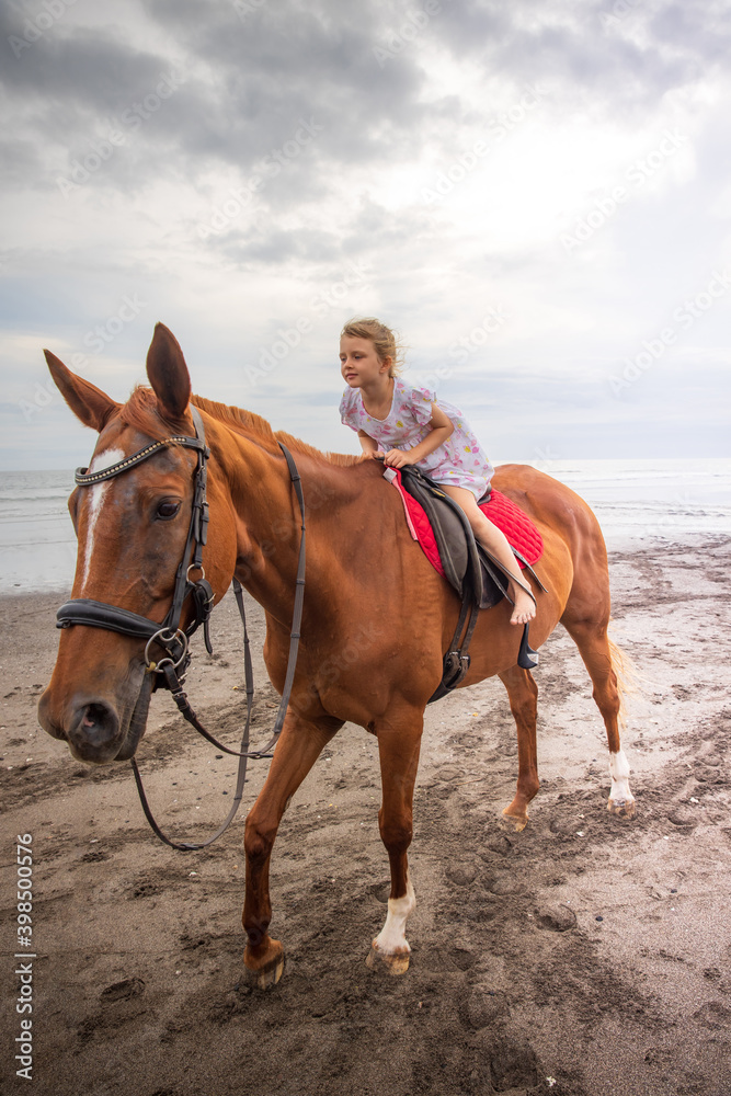 Pretty little girl riding horse on the beach. Happy childhood. Sunset time by the sea. Outdoor activities. Vacation concept. Bali, Indonesia Pretty little girl riding horse on the beach. Happy childhood. Sunset time by the sea. Outdoor activities. Vacation concept. Bali, Indonesia