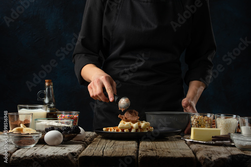 The professional chef pours ice-cream ball for serving waffles on rustic wooden table with ingredients on dark blue background. Backstage of cooking sweet dessert for breakfast. Frozen motion.