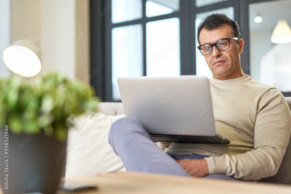 Freelance worker. Portrait of latin middle aged business man with eyeglasses working at home on some project, sitting on a sofa with his laptop Freelance worker. Portrait of latin middle aged business man with eyeglasses working at home on some project, sitting on a sofa with his laptop