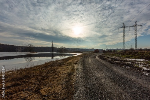 Rural landscape with dark brown gravel road between lake and a field with high-voltage power lines. Cloudy winter evening with blue sky and sun hiding behind grey clouds.