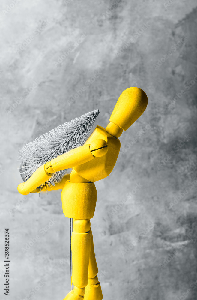Yellow wooden mannequin is carrying tiny christmas tree, concept of preparation for Xmas holidays, going home for festive time concept, trendy colors of the year 2021 with gray background