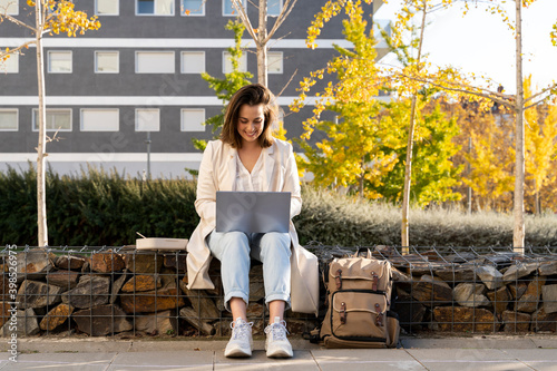 Smiling businesswoman using laptop while sitting on retaining wall in city