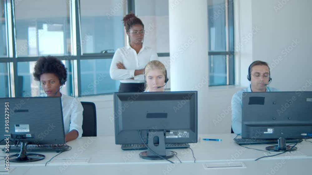 Strict female boss watching call center employees in headsets working at computers. Front view. Female business leader or telemarketing concept