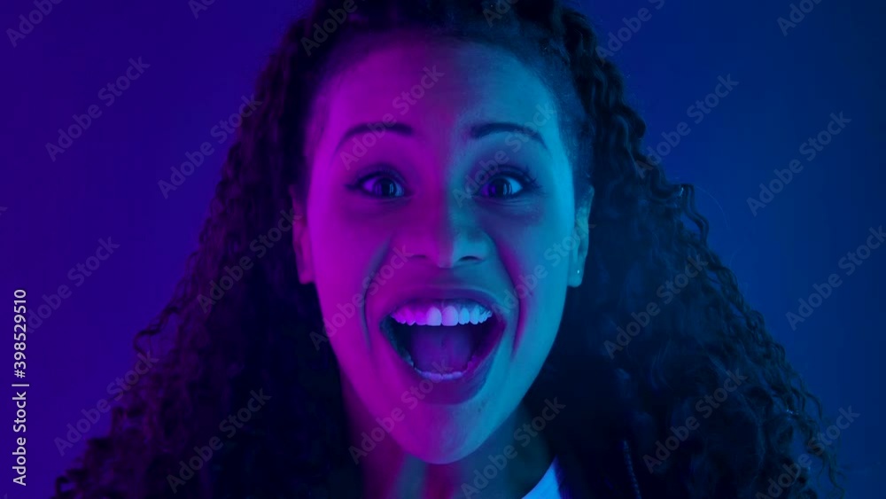 Portrait of a young lovely female African American looking at the camera with shocked and surprised wow face expression. Close up face illuminated with purple and blue neon lights. Slow motion.