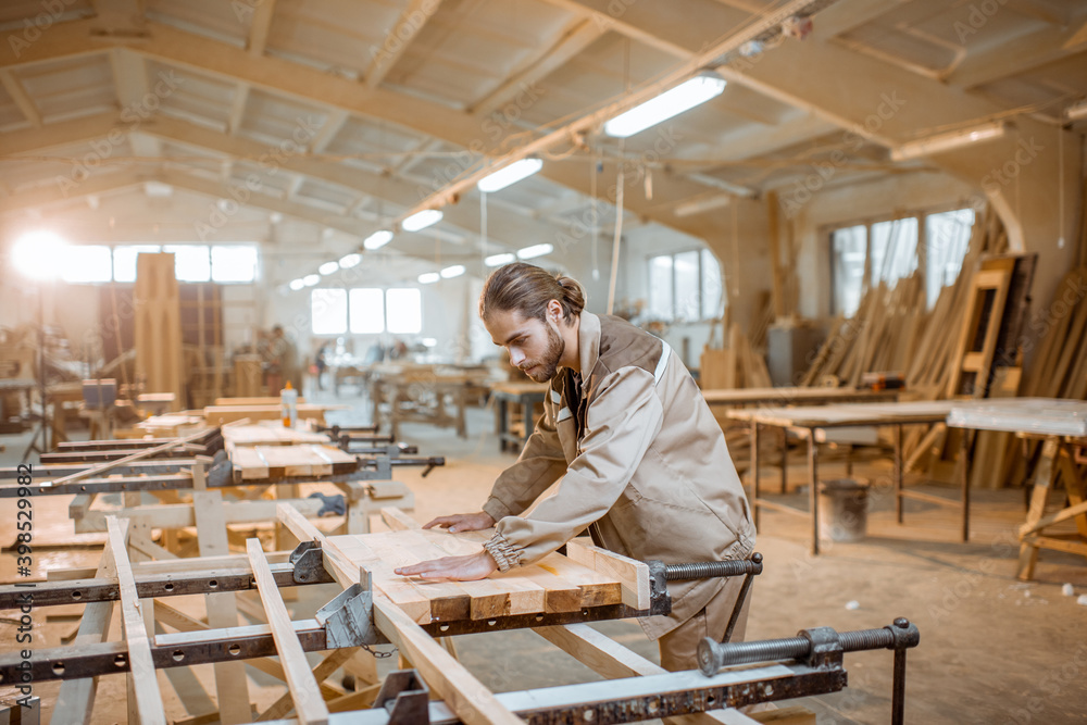 Handsome carpenter in uniform gluing wooden bars with hand pressures at the carpentry manufacturing