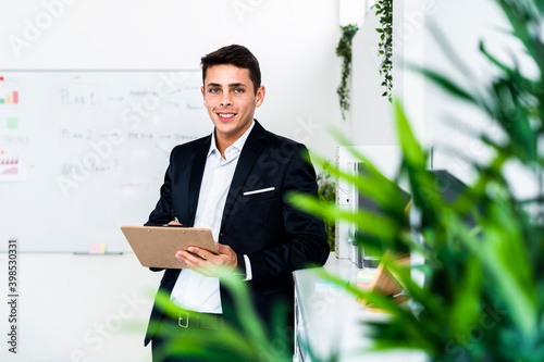 Smiling male professional writing on clipboard while standing by cabinet at office
