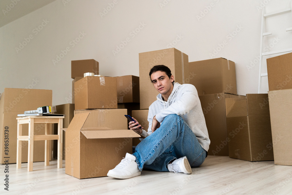Young man using mobile phone while sitting against cardboard boxes in new house Young man using mobile phone while sitting against cardboard boxes in new house