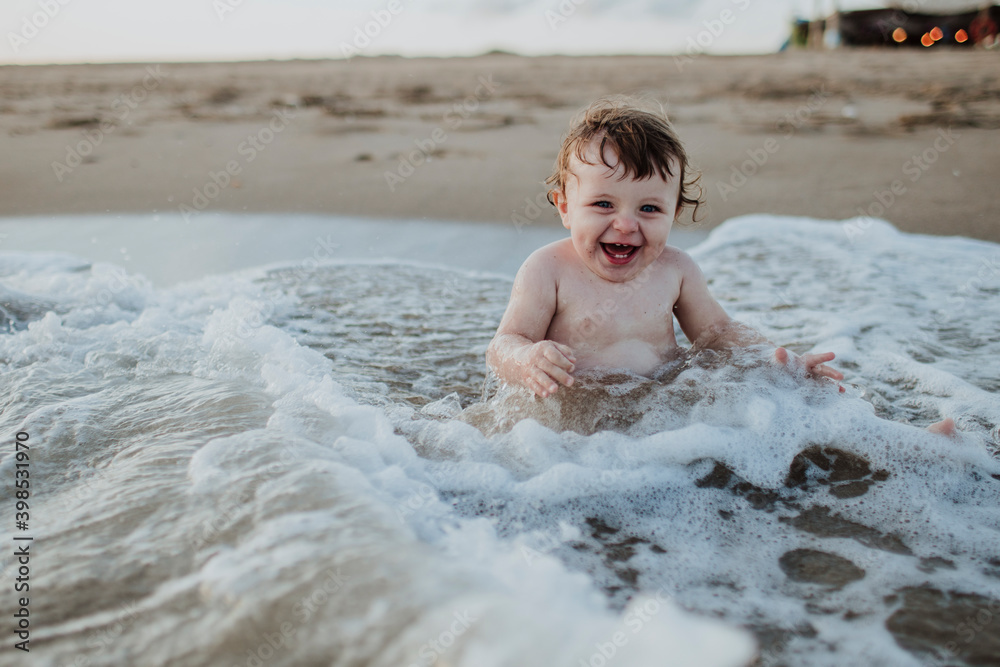 Male toddler laughing while sitting in water at beach during sunset