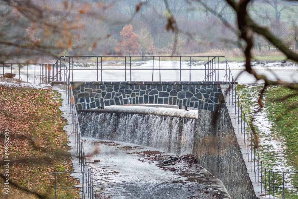 Obraz premium A lake embankment with outlet, a stone bridge with railing. White snow on green grass. Branch with dry leaves framing the image.