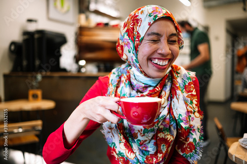 Woman in floral hijab laughing while holding coffee cup at cafe photo