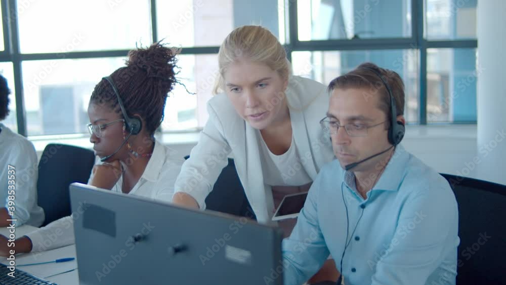 Young manager helping call center operators with new software, pointing at monitors and speaking. Customers assistance or telemarketing concept.