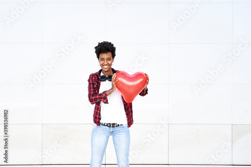 Happy young female hipster holding red heart shape balloon against white wall photo