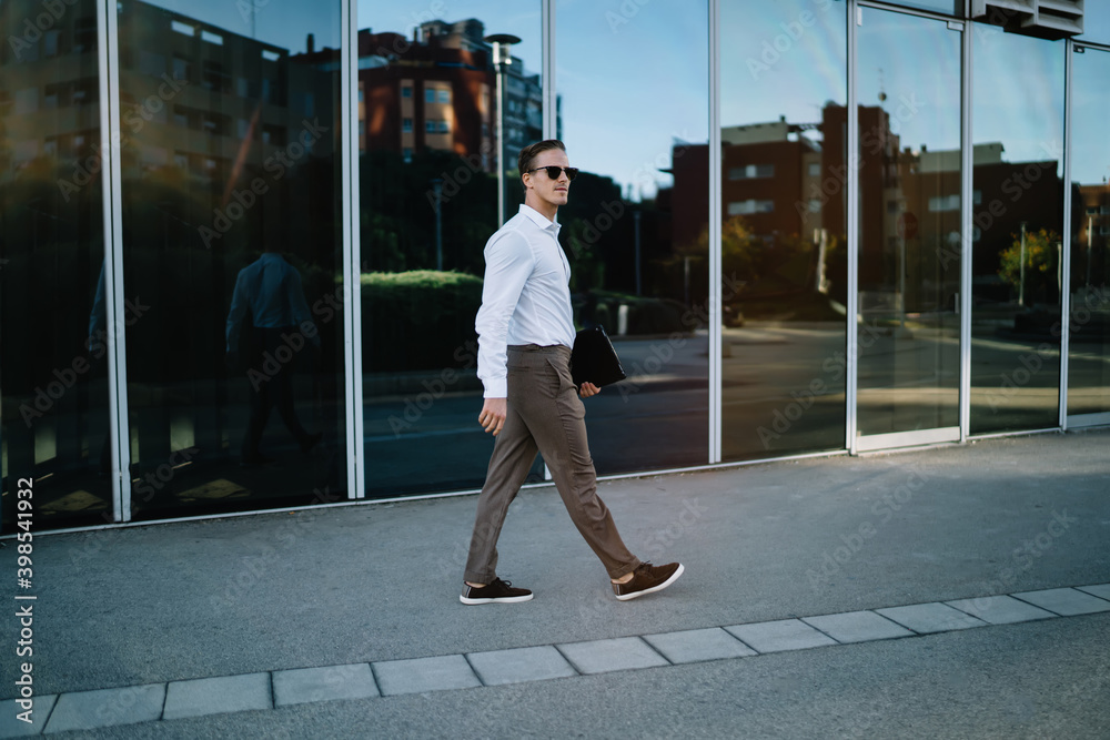 Side view of confident businessman in stylish sunglasses and smart casual clothing walking at urban street getting to corporate meeting in financial district, Caucasian male manager in trendy outfit
