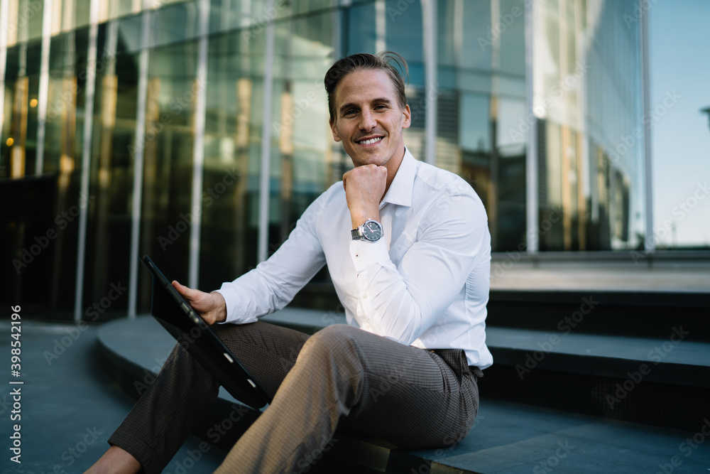 Portrait of cheerful male employee dressed in stylish smart casual apparel sitting at street stairs and smiling at camera, happy businessman with modern touch pad in hand posing in city downtown Portrait of cheerful male employee dressed in stylish smart casual apparel sitting at street stairs and smiling at camera, happy businessman with modern touch pad in hand posing in city downtown