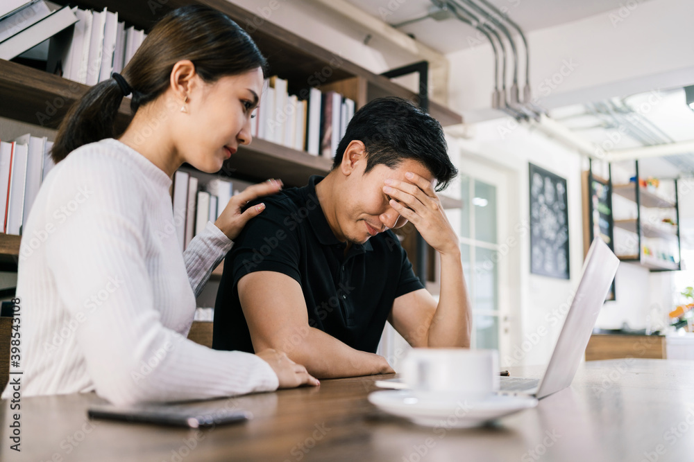 The sad unhappy handsome man sitting in front of a laptop, holding his forehead while having a headache with a woman cheering up by touching his shoulder. The sad unhappy handsome man sitting in front of a laptop, holding his forehead while having a headache with a woman cheering up by touching his shoulder.