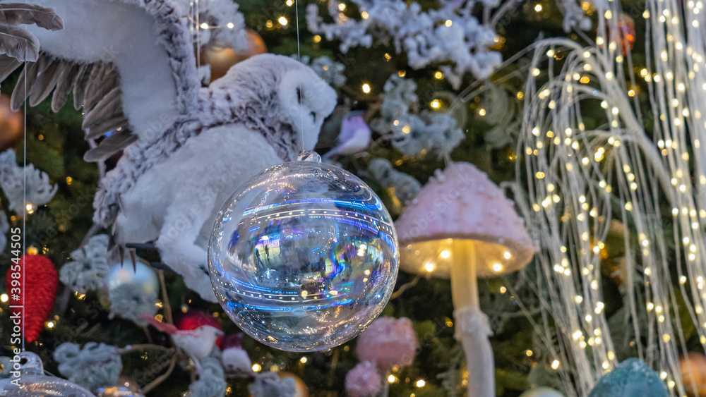Close-up of toy in the form of flying owl on spruce branch. In the foreground is a large Christmas ball. Decorated christmas tree background. Happy new year concept. Blurred background with lights.