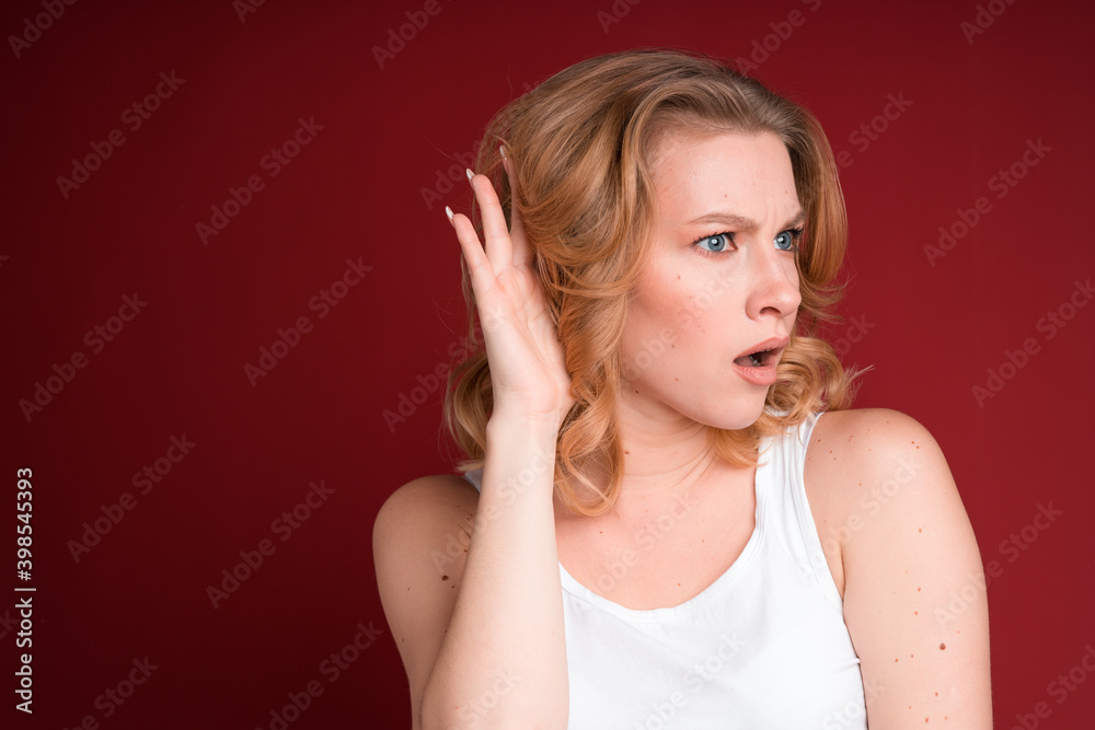 Blond woman with curly hair put her hand to ear in white tank top isolated on red background. Blond woman with curly hair put her hand to ear in white tank top isolated on red background.