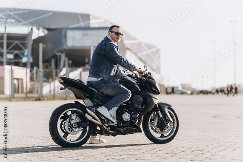 A man in denim clothes sitting on a black sports motorcycle against the backdrop of a large modern building