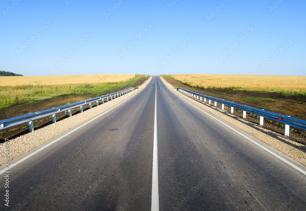 Fototapeta premium New asphalt road surface on the background of green and yellow fields to the horizon. The construction site for the road works. Highway on the background of a rural landscape 