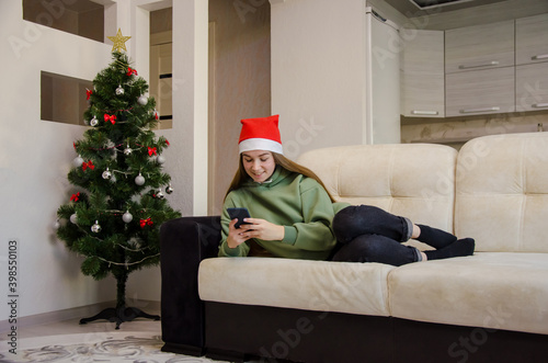 the girl lies on the couch and writes a message to friends and colleagues on the phone in the background a Christmas tree. girl wishes happy holidays online © Valiantsina