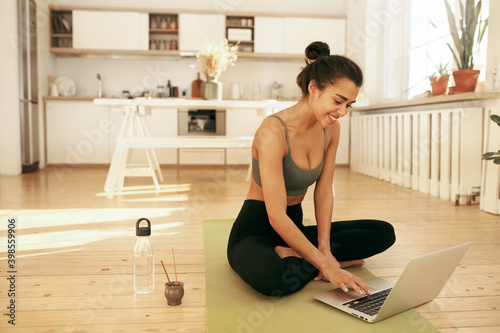 Technology and active lifestyle concept. Beautiful young slim Hispanic woman enjoying yoga practice early in the morning, sitting in stylish interior, playing music for meditation on laptop