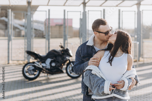 Sexy girl and guy hugging and kissing on the background of a sports motorcycle standing in the parking lot near a large football stadium