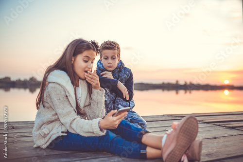 Two little friends, boy and girl watching something interesting on smartphone sitting by the lake in the evening