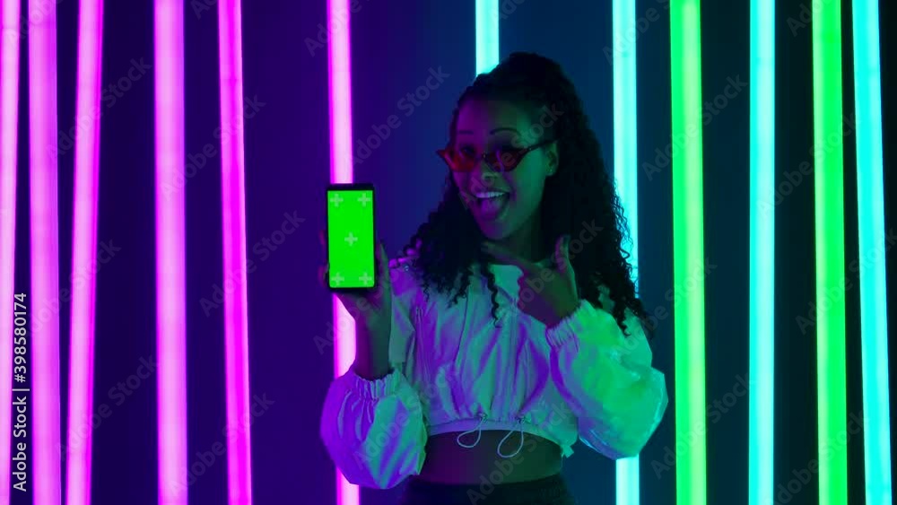 Portrait of a young lovely female African American in stylish sunglasses points a finger at a smartphone with a green screen chroma key. Slow motion. Portrait of a young lovely female African American in stylish sunglasses points a finger at a smartphone with a green screen chroma key. Slow motion.