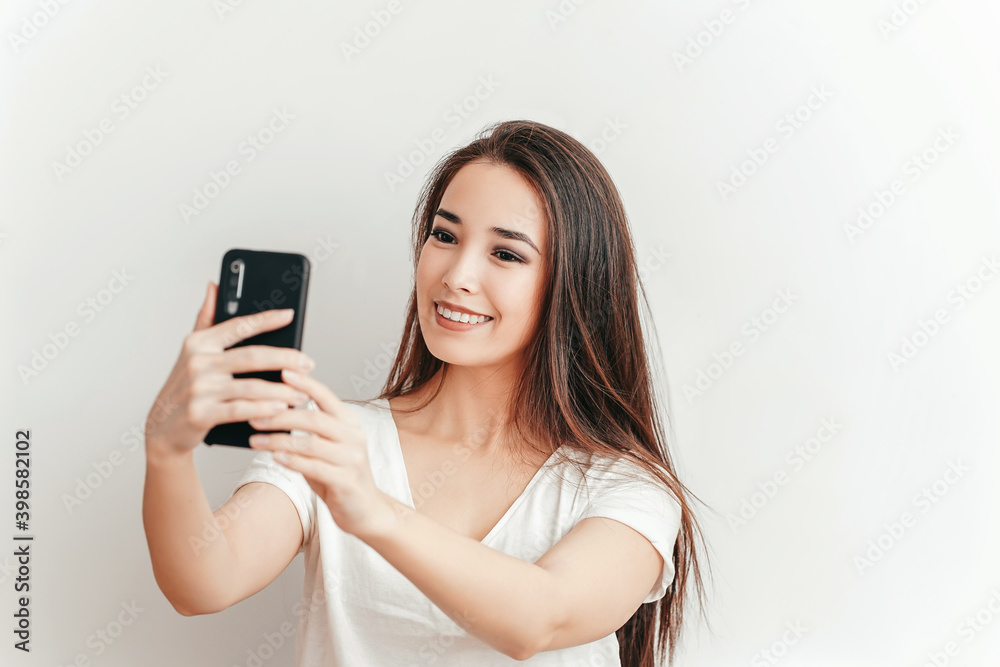 Beautiful Asian smiling girl takes selfie on mobile phone on white background in white T-shirt with beautiful smile Beautiful Asian smiling girl takes selfie on mobile phone on white background in white T-shirt with beautiful smile