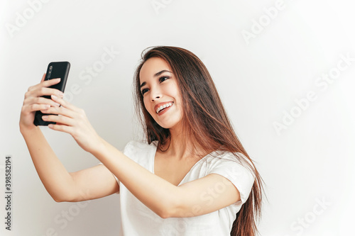 Beautiful Asian smiling girl takes selfie on mobile phone on white background in white T-shirt with beautiful smile