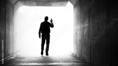 фотографія A single male in a dark tunnel exiting and walking towards a bright white light while waving goodbye or hello