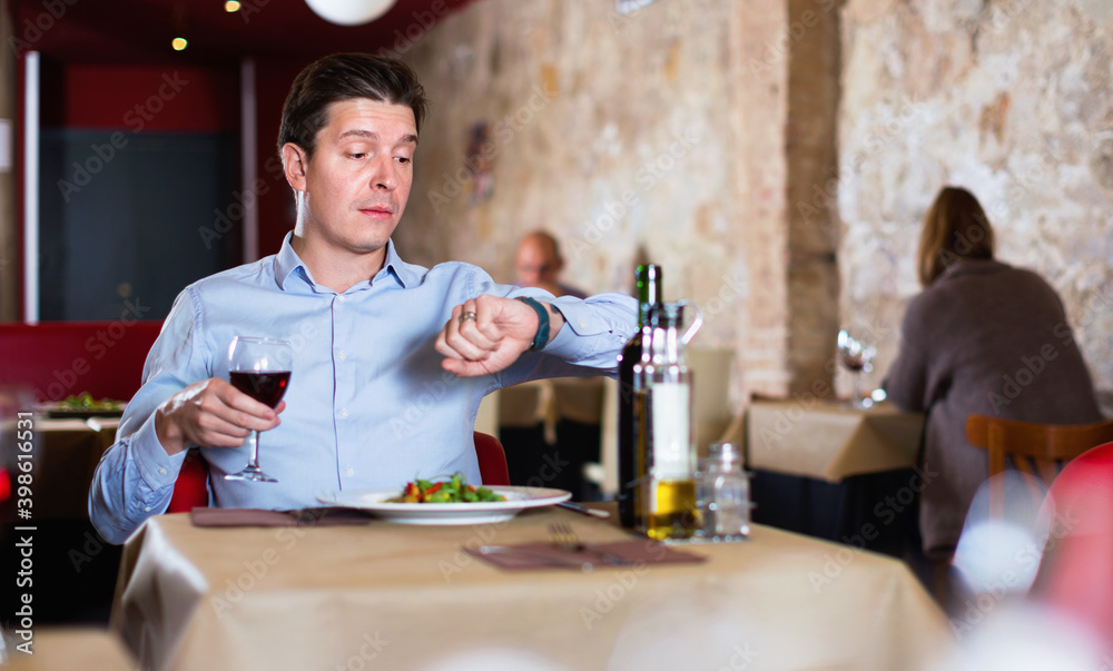Young positive man sitting in restaurant with wine glass, looking at clock in anticipation of friend