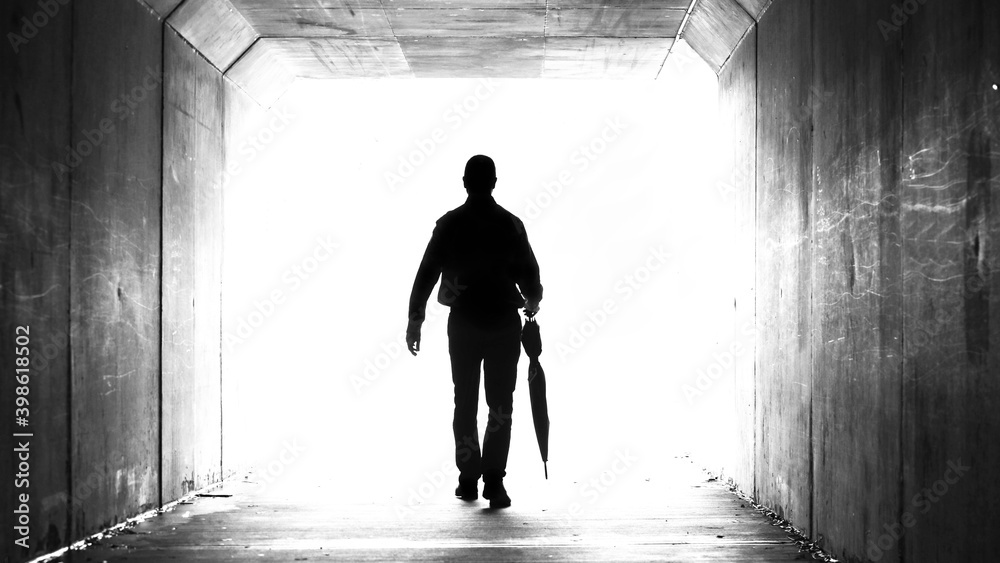 Obraz premium A silhouette of a man holding a folded up umbrella leaving a dark tunnel walking towards the light. Weather, climate, leaving and departure theme
