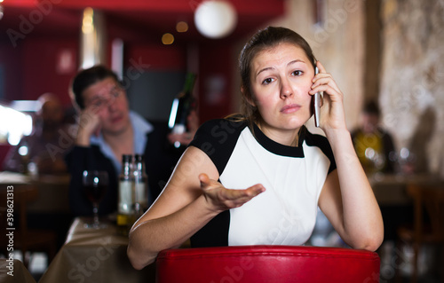 Unhappy young female talking on mobile phone sitting apart in restaurant with drunk man behind