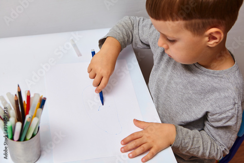 Cute little boy with blond hair draws colored pencils at home. Draws at the white table.