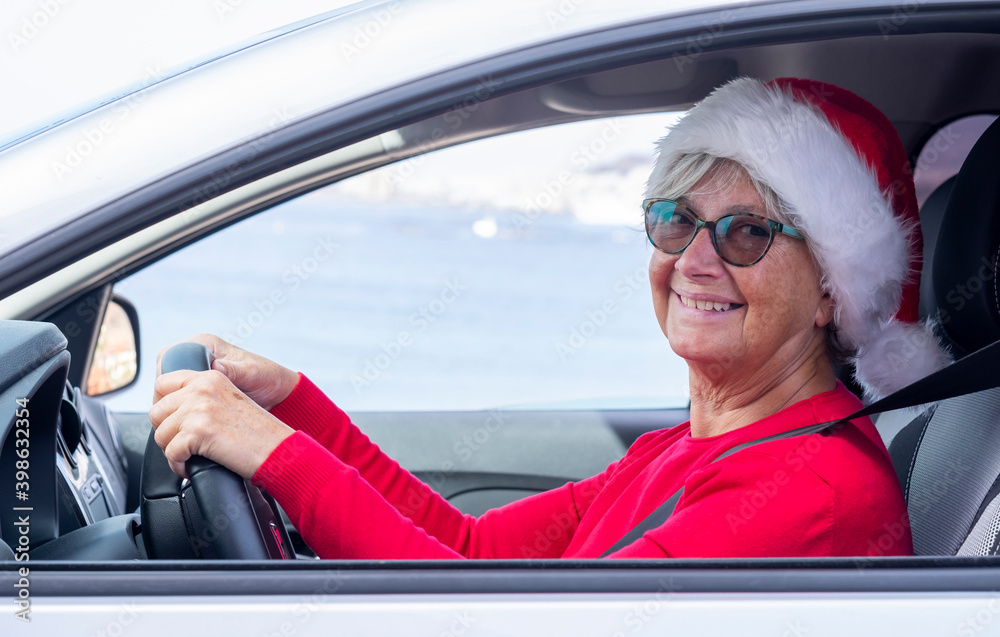 Smiling senior woman red dressed wearing a Christmas hat smiles driving the car looking at camera. Smiling senior woman red dressed wearing a Christmas hat smiles driving the car looking at camera.