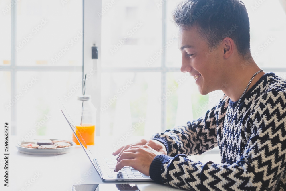 Handsome, smiling teenager using laptop at home in front of the window - takes a break with cookies and orange juice. Millenial generation enjoying technology and social Handsome, smiling teenager using laptop at home in front of the window - takes a break with cookies and orange juice. Millenial generation enjoying technology and social