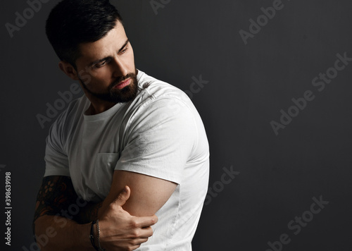 фотографія Handsome modern brutal bearded muscular man in t-shirt and with tattoo on arms stands side to camera looking back at copy space over grey background