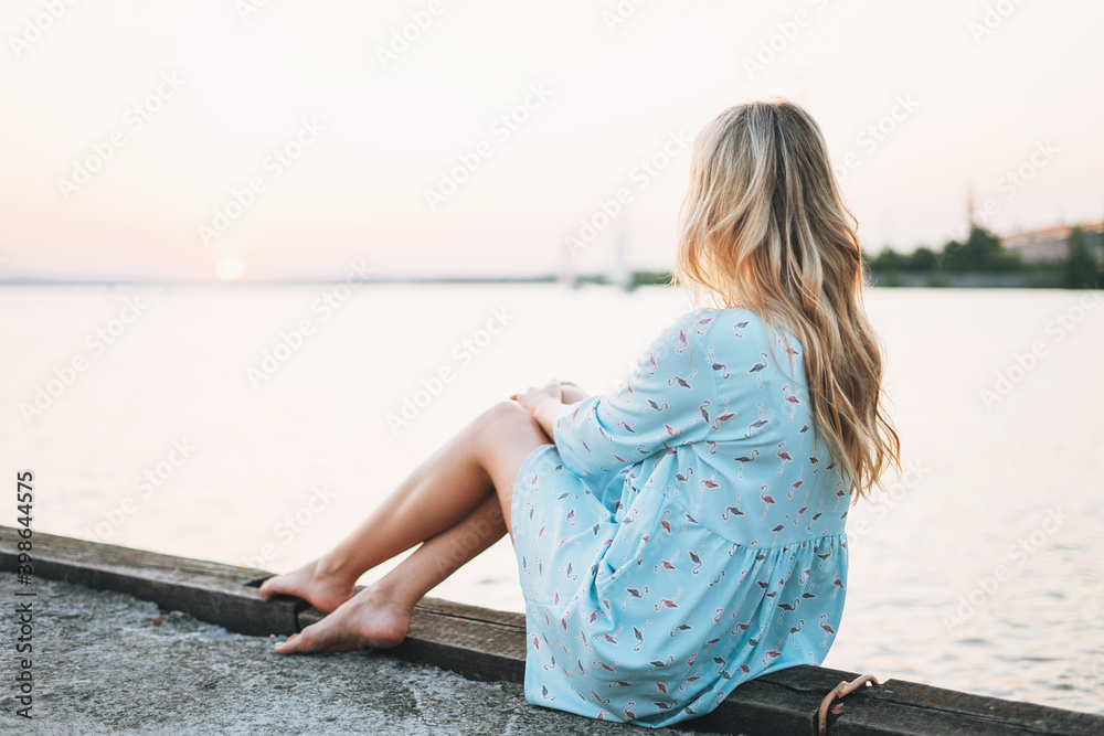 Beautiful blonde young woman in blue dress sitting on pier and looking on sunset