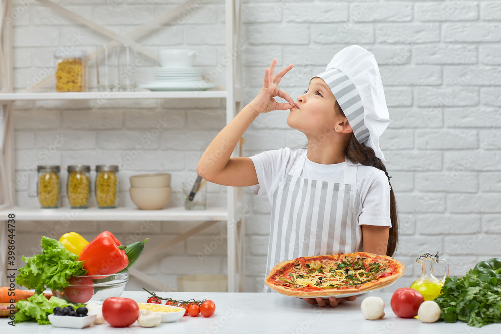 child making tasty delicious gesture by kissing fingers. little girl in chef hat and an apron cooking pizza in the kitchen. child making tasty delicious gesture by kissing fingers. little girl in chef hat and an apron cooking pizza in the kitchen.
