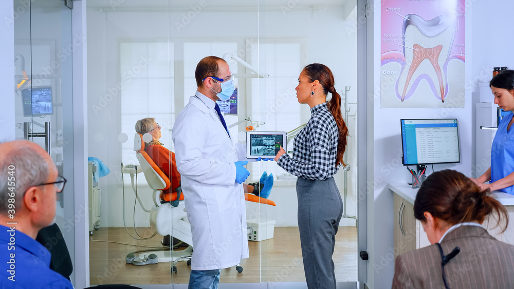 Orthodontist using tablet explaining dental x ray to patient standing in waiting area of stomatologic office. Dentist showing to woman radiography using digital device working in modern clinic. Orthodontist using tablet explaining dental x ray to patient standing in waiting area of stomatologic office. Dentist showing to woman radiography using digital device working in modern clinic.