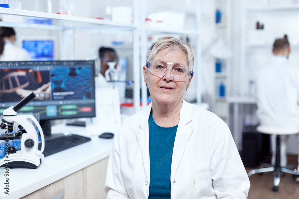Obraz premium Senior medical research in scientific clinic with virus on computer display. Elderly scientist wearing lab coat working to develop a new medical vacine with african assistant in the background.