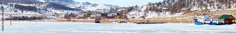 Obraz premium Winter Baikal Lake. A panoramic view of the coast of the Kurkut Bay - a popular tourist destination with wooden houses, a ship on a frozen pier and an exit to the ice road to Olkhon Island
