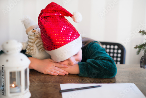 young blond girl in Santa hat falls asleep on the table while writing a wish letter list to Santa