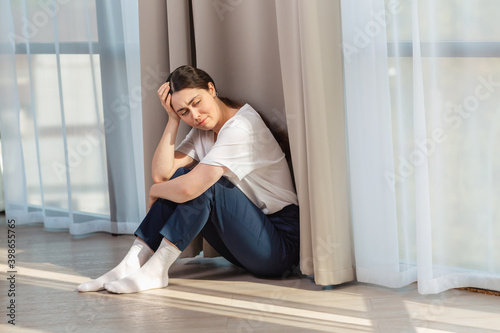 Depressed crying woman holding her head with her hand and sitting on the floor. Copy space. Windows on the background. The concept of domestic violence