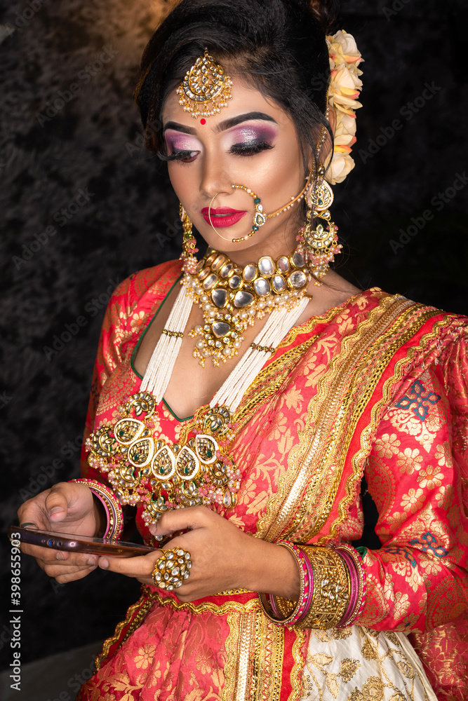 Portrait of very beautiful young Indian bride in luxurious bridal costume with makeup and heavy jewellery looking at mobile. Wedding fashion.