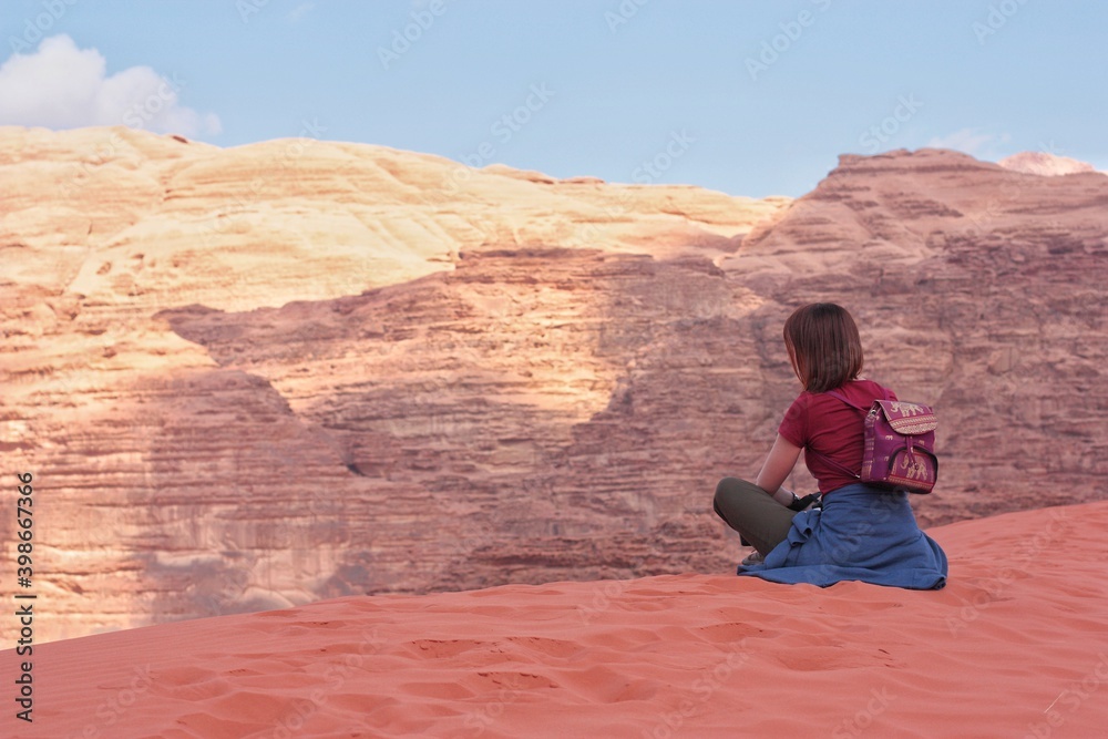 a girl sits on the edge of a sand dune in front of a relief mountain, nature jordan