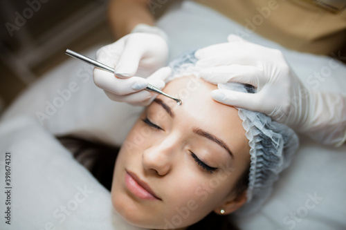 Fotografija Young beautiful woman is lying on a medical couch while a doctor cosmetologist in a white gloves is doing a cleaning therapy named facial mechanical cleaning for her face