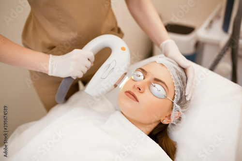 Young beautiful woman on beauty cosmetology procedure in spa salon. Therapist beautician makes a laser treatment to young woman's face at beauty SPA clinic. Facial laser hair removal epilation